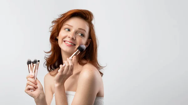 Rood harige jonge vrouw houdt cosmetische borstels in de buurt van gezicht, grijze achtergrond — Stockfoto
