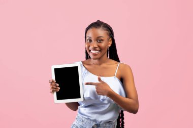 Pozitif genç Afro kadın pembe arka planda boş ekranı olan tablet bilgisayarına işaret ediyor, web sitesi veya uygulama için modelleme yapıyor