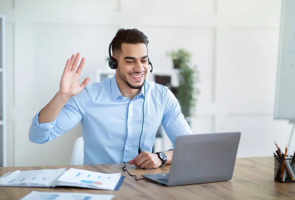 Sonriente chico árabe en auriculares hablando con su socio de negocios, tener videollamada en línea, saludando a la cámara en la oficina —  Fotos de Stock