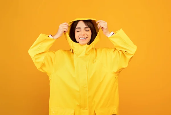 Excitada senhora milenar em capa impermeável amarela vestindo capuz, de pé sobre fundo amarelo e sorrindo — Fotografia de Stock