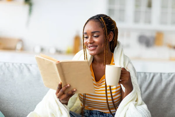 Radosna czarna kobieta pije herbatę i czyta książkę w domu — Zdjęcie stockowe