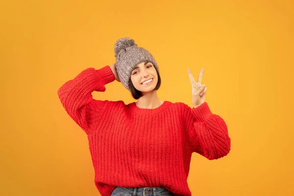 Jovem senhora feliz em chapéu de inverno tricotado mostrando vitória ou paz mão sinal gesto, posando sobre fundo laranja — Fotografia de Stock