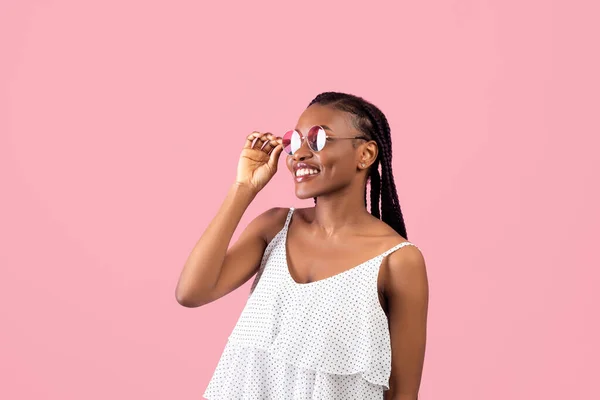 Retrato de una hermosa mujer negra en vestido y gafas de sol mirando a un lado en el fondo del estudio rosa — Foto de Stock