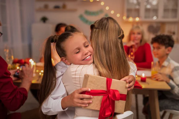 Happy little girl hugging her mother, receiving gift box during festive dinner at home — Stock Photo, Image
