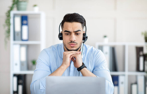 Worried millennial Arab man in headphones making online video call with business partner, having problem at office