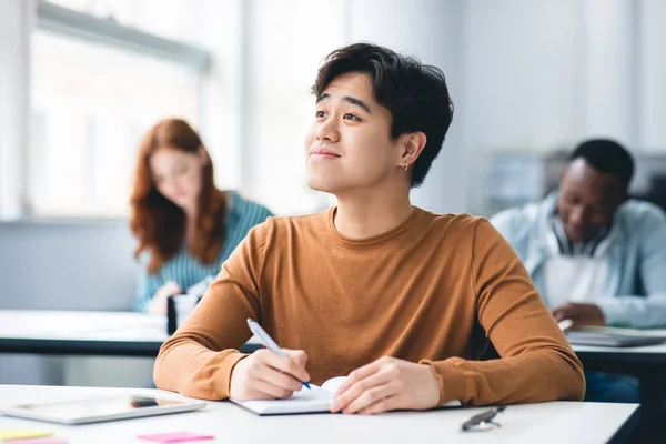 Sourire asiatique étudiant assis à bureau dans salle de classe regardant loin — Photo