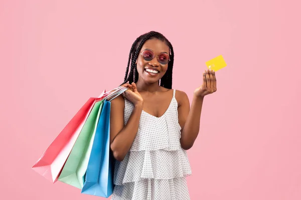Легкий шопинг. Радостная афро-леди держит кредитную карту и яркие сумки покупателей на розовом фоне студии — стоковое фото