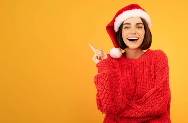 Noel indirimi konsepti. Noel Baba şapkalı neşeli kadın sarı stüdyo arka planında boş bir yeri işaret ediyor. — Stok fotoğraf