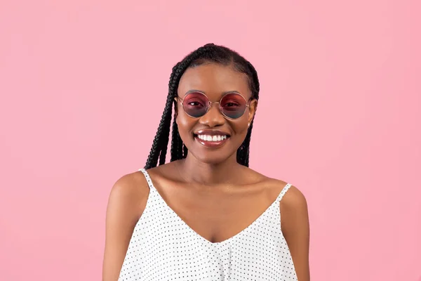 Portret chłodnej czarnej damy w afro w stylowej sukience i okularach przeciwsłonecznych, uśmiechniętej do kamery na różowym tle — Zdjęcie stockowe