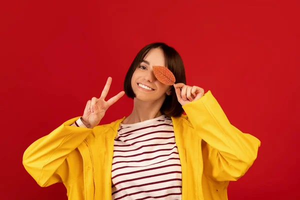 Mujer excitada cerrando el ojo con hoja de otoño y mostrando gesto de paz, sonriendo a la cámara sobre fondo rojo — Foto de Stock