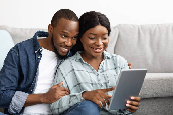 Black Happy Millennial Couple Using Digital Tablet Together While Relaxing At Home