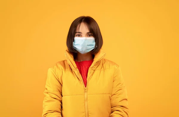 Coronavirus concept. Jonge vrouw in beschermende gezichtsmasker en winter jas poseren over gele achtergrond — Stockfoto