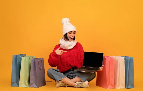 Mujer joven en traje cálido que muestra la pantalla vacía del ordenador portátil, señalando con el dedo en él y sentado entre las bolsas de compras de colores — Foto de Stock