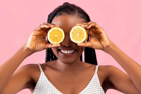 Vitaminas para beleza e cuidados com a pele. Mulher afro positiva cobrindo ambos os olhos com metades de limão no fundo rosa — Fotografia de Stock