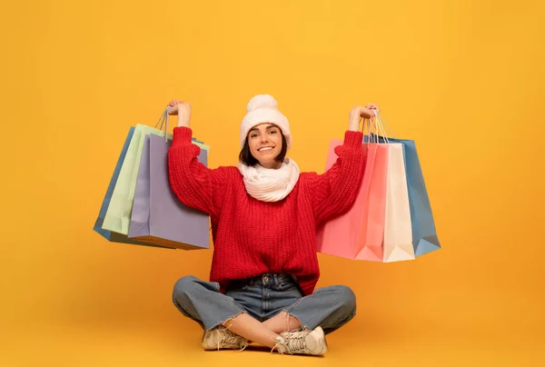 Velký zimní výprodej. vzrušená mladá žena v bílém klobouku a pletený svetr s nákupními taškami, sedí na žlutém pozadí — Stock fotografie