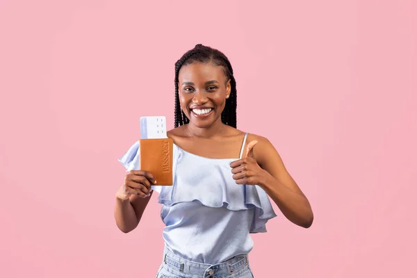 Happy black female traveller segurando passaporte com bilhete de embarque de avião e mostrando o polegar no fundo rosa — Fotografia de Stock