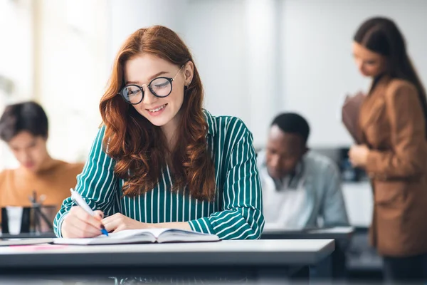 Señora sonriente sentada en el escritorio en el aula escribiendo en cuaderno — Foto de Stock