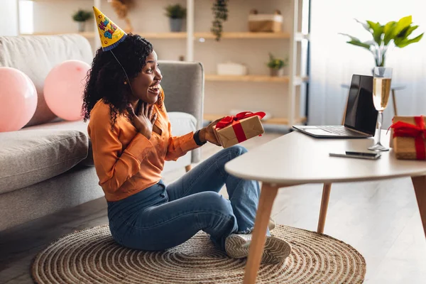 Surprised African Lady Holding Birthday Gift At Laptop Indoor, Side-View — Stock Photo, Image