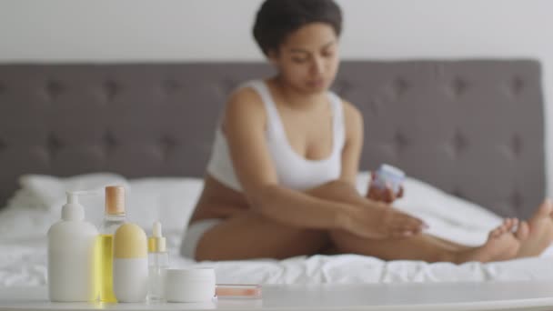 Mujer afroamericana borrosa aplicando crema hidratante en las piernas piel sentada en la cama, botellas de cosméticos en primer plano — Vídeos de Stock