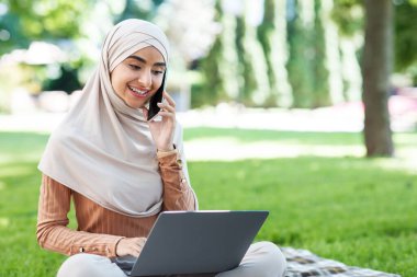 Tesettürlü genç ve çekici bir ortadoğulu kadın bilgisayarına bakıyor, telefonla arıyor, uzaktan parkta çalışıyor.
