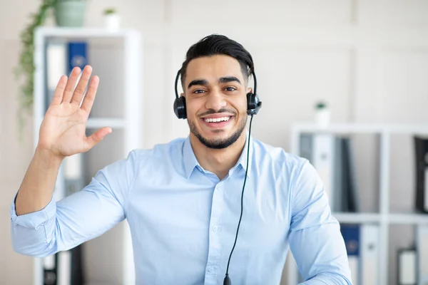 Communicatie op afstand. Happy Arab guy met headset met video call op kantoor, zwaaien naar webcam — Stockfoto