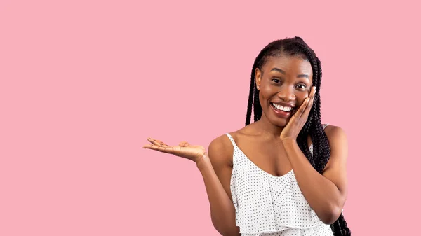 Возбужденная афро женщина в стильном платье трогает ее лицо, показывая что-то на пустом пространстве на розовом фоне — стоковое фото