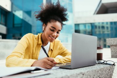 Gülümseyen siyahi kız laptopta notlar alıyor. Dışarıda öğreniyor.