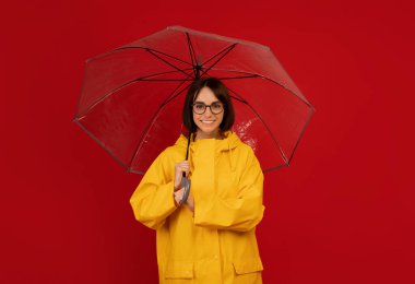 Yağmurlu bir günde kendimi güvende hissediyorum. Kırmızı stüdyo arka planında şeffaf şemsiye altında duran gülümseyen kadının portresi