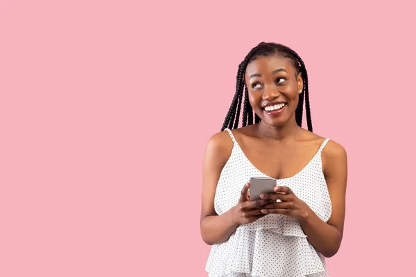 Dreamy black woman using smartphone, looking aside at free space on pink studio background — Stok Foto