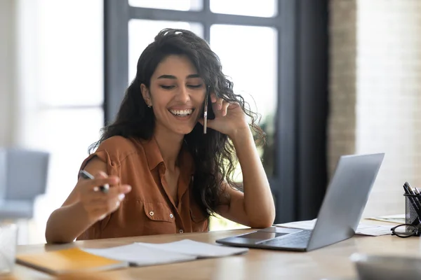 Jeune femme d'affaires parlant sur un téléphone portable et travaillant sur un ordinateur portable à partir de son bureau à domicile, discutant de problèmes de travail à distance — Photo