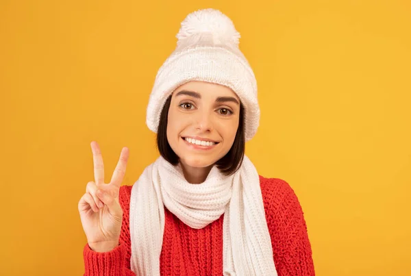Happy lady in warm sweater and knitted hat smiling and making peace gesture on yellow studio background — Stock Photo, Image