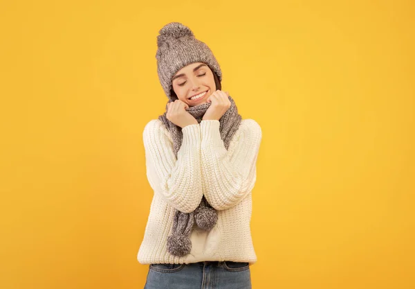 Senhora bonita feliz vestindo camisola de malha branca, cachecol cinza e chapéu, de pé com os olhos fechados sobre o fundo do estúdio amarelo — Fotografia de Stock