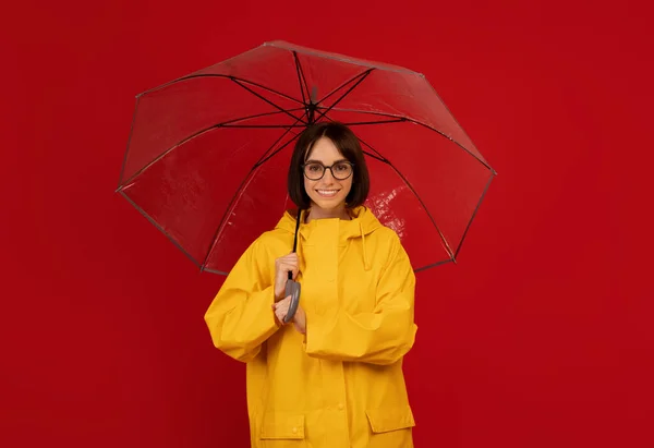 Νιώθω προστατευμένη τις βροχερές μέρες. Πορτρέτο της χαμογελαστή κυρία στέκεται κάτω από διαφανή ομπρέλα πάνω από το κόκκινο φόντο στούντιο — Φωτογραφία Αρχείου