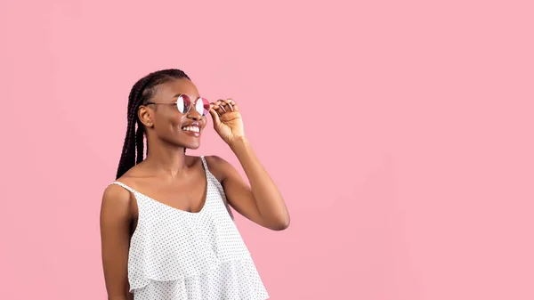 Mulher afro adorável na roupa da moda tocando óculos de sol, olhando para o espaço vazio no fundo rosa, design de banner — Fotografia de Stock