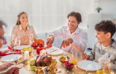 Cheerful young man celebrating Christmas or Thanksgiving at home, cutting roasted turkey for festive family dinner clipart