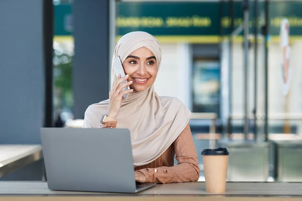 Elfoglalt mosolygós vonzó fiatal arab hölgy hidzsáb hívások telefonon, ül laptop és kávé kávézóban — Stock Fotó