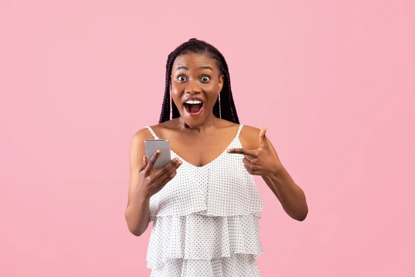 ショックを受けた黒の女性は興奮してスマートフォンを指して、宝くじやオンラインカジノベットでピンクの背景 — ストック写真
