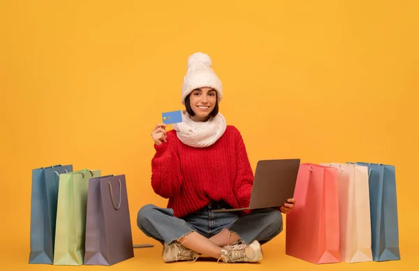 Fácil de comprar. Mujer feliz en ropa de punto usando el ordenador portátil y la tarjeta de crédito para las compras en línea, fondo amarillo — Foto de Stock