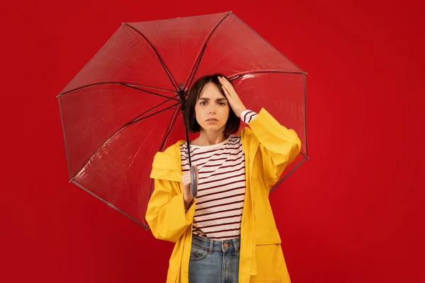 Begreppet väderberoende. ung kvinna vidrör huvudet och står under paraply, poserar över röd bakgrund — Stockfoto