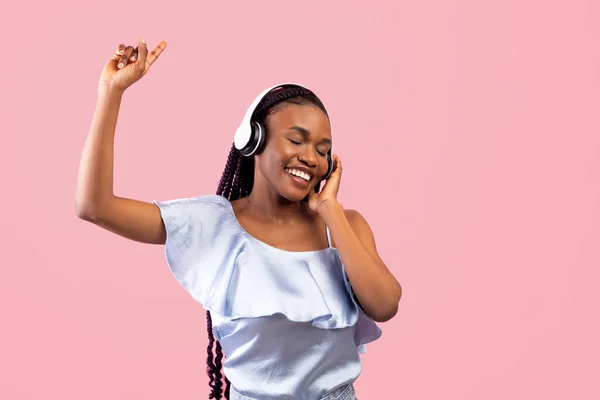 Mujer negra positiva en auriculares inalámbricos bailando con música popular sobre fondo de estudio rosa — Foto de Stock