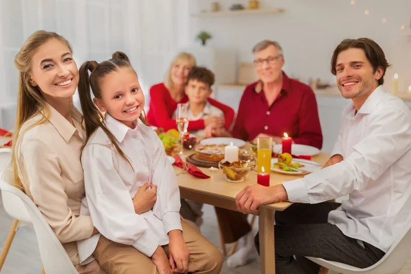 Porträt einer glücklichen Mehrgenerationenfamilie, die am festlichen Tisch sitzt, in die Kamera blickt und lächelt — Stockfoto