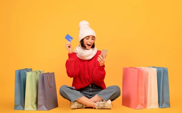 Erleichtern Sie Ihren Einkauf. Überglückliche Dame in Strickmütze mit Smartphone und Kreditkarte sitzt zwischen Einkaufstaschen — Stockfoto