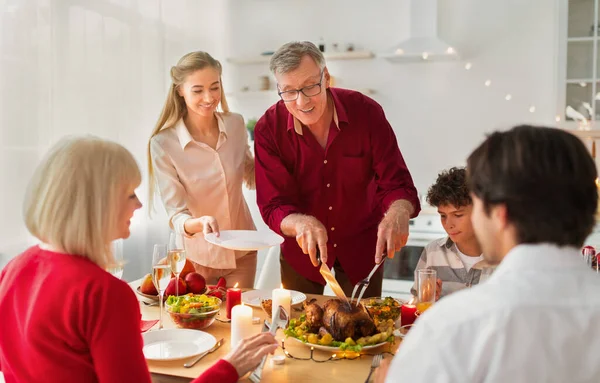 Boldog idős férfi ünnepi sült pulykát szeletel, otthon ünnepli a hálaadást vagy a karácsonyt a családjával. — Stock Fotó