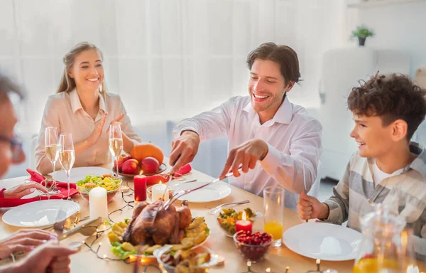 Cheerful young man celebrating Christmas or Thanksgiving at home, cutting roasted turkey for festive family dinner — Stock Photo, Image