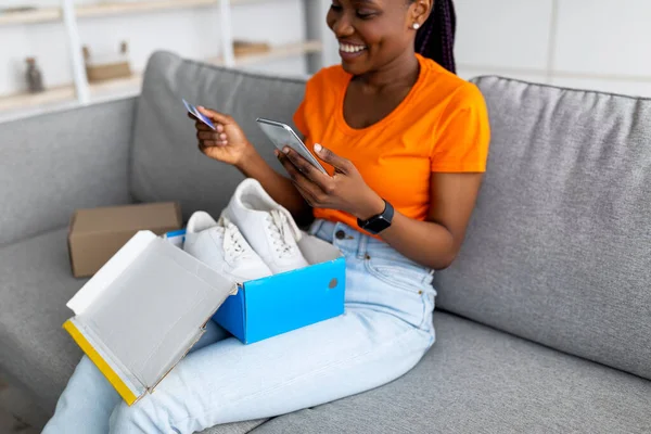 Svart kvinna packa upp paket med nya sportskor, hålla kreditkort och smartphone, skriva online-butik feedback — Stockfoto