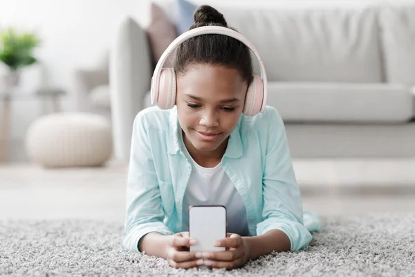 Concentrato sorridente adolescente afroamericano ragazza che studia, guardando lezione online sul telefono, si trova sul pavimento — Foto Stock