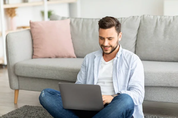Mosolygó Y-generációs férfi dolgozik otthonról, ül a padlón, és használja laptop, szabad hely — Stock Fotó