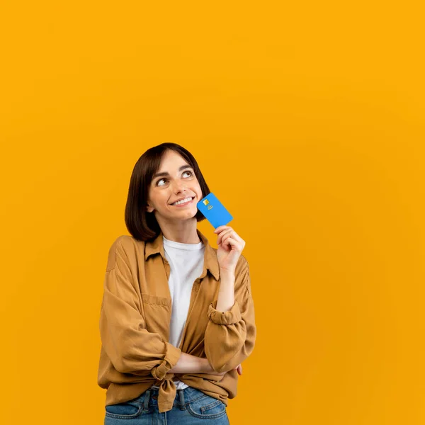 Time for shopping. Pensive woman holding credit card and looking away, planning what to buy, yellow background, crop — Stock Photo, Image