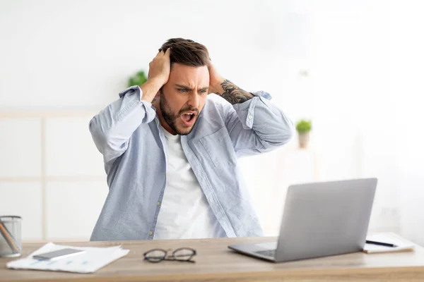 Rozzlobený muž se softwarem, webové stránky chyba, při pohledu na obrazovku notebooku a gestikulace, sedí na pracovišti v interiéru domu — Stock fotografie