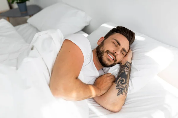 Счастливого утра. Удовлетворенный парень просыпается, чувствуя себя отдохнувшим, счастливым бородатым черным парнем, лежащим на простынях из льна — стоковое фото